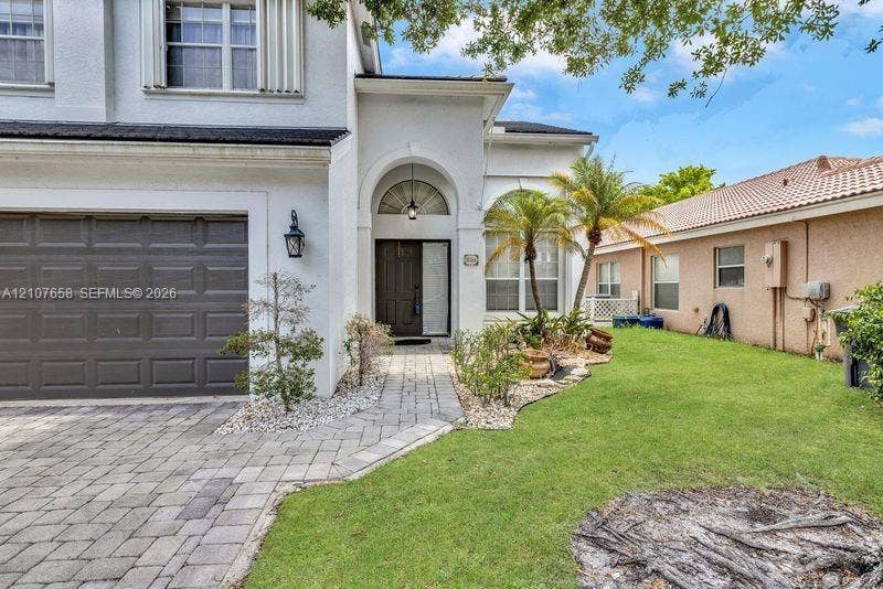 Stucco exterior featuring an arched entryway, paver driveway, dark-toned garage door, and a manicured lawn with mature palm landscaping