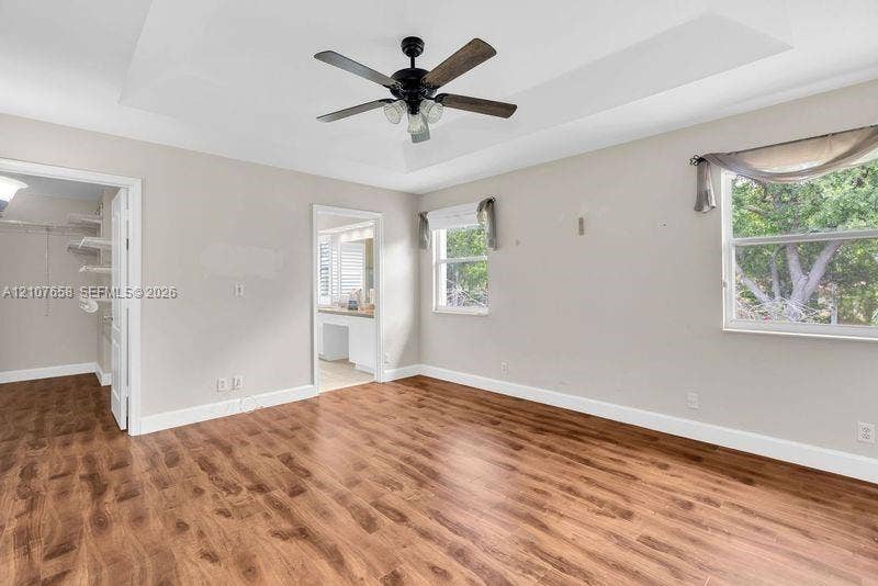 Tray ceiling featuring a central multi-blade ceiling fan with integrated lighting, wood-finish flooring, and dual windows