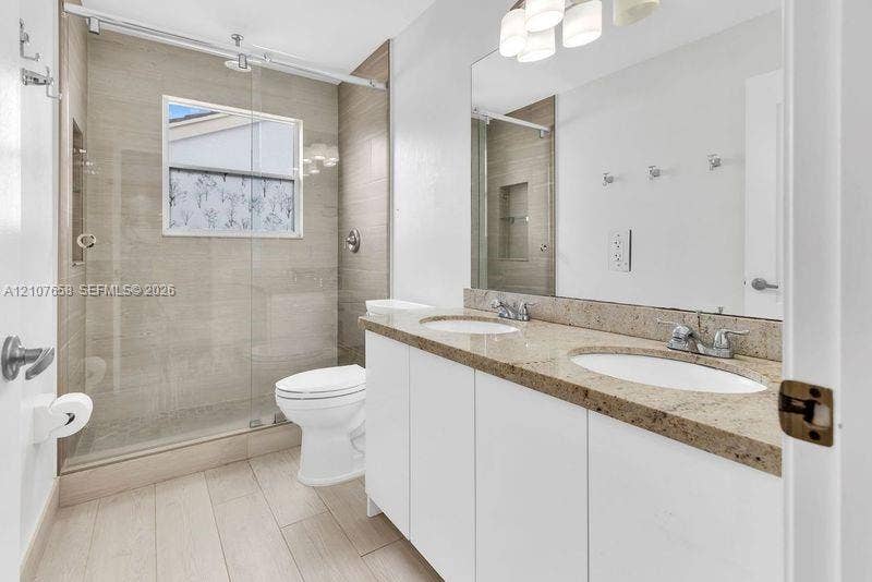 Dual-sink vanity with stone countertop, walk-in shower featuring glass sliding doors and tiled surround, and wood-finish flooring