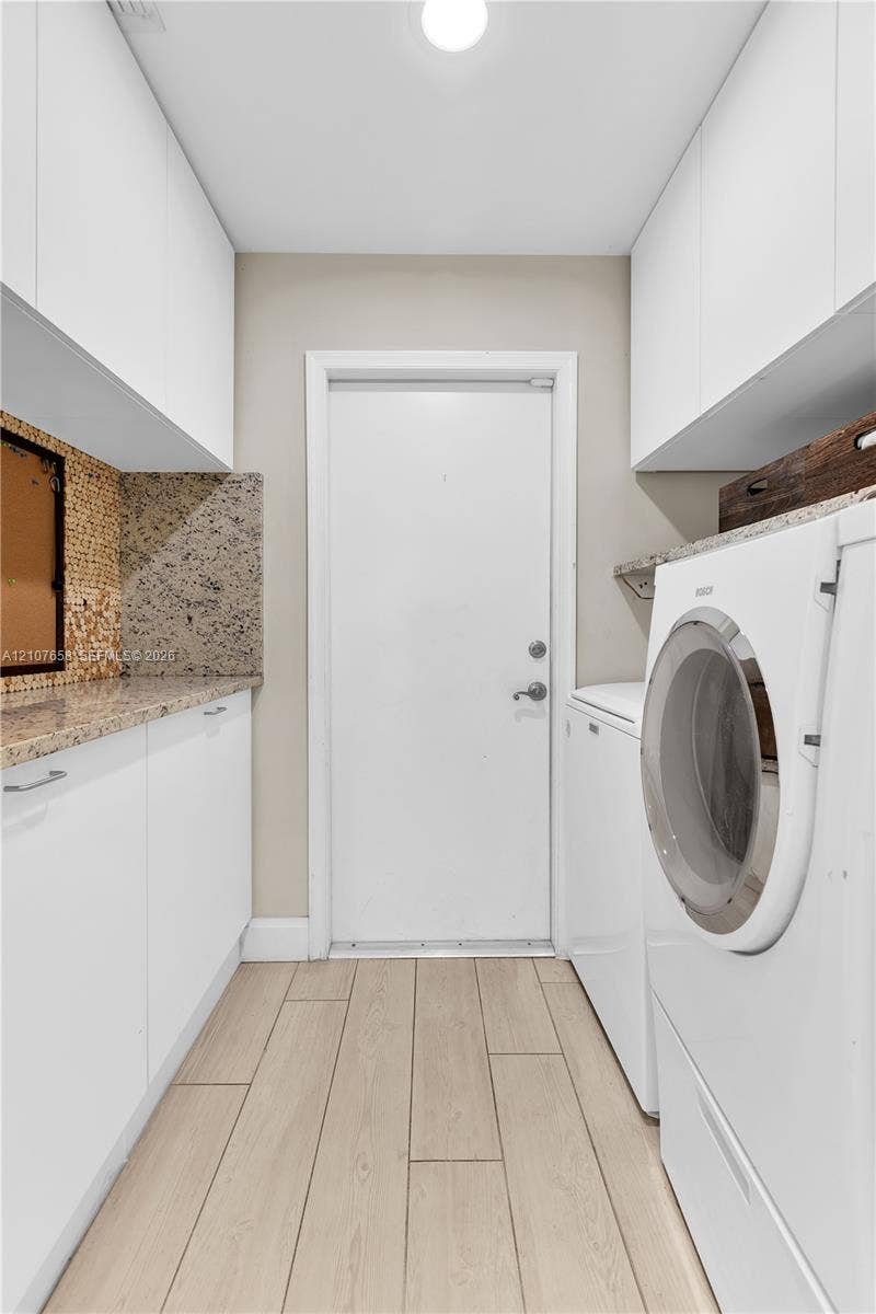 Utility room featuring wood-finish flooring, white flat-panel upper and lower cabinetry, stone-finish countertops, and a decorative mosaic tile backsplash
