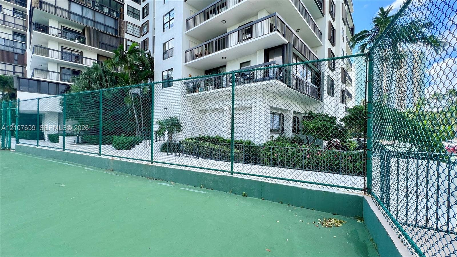 Recreational court with a high-tension perimeter fence, situated on the grounds of a multi-story building featuring private balconies and landscaped walkways