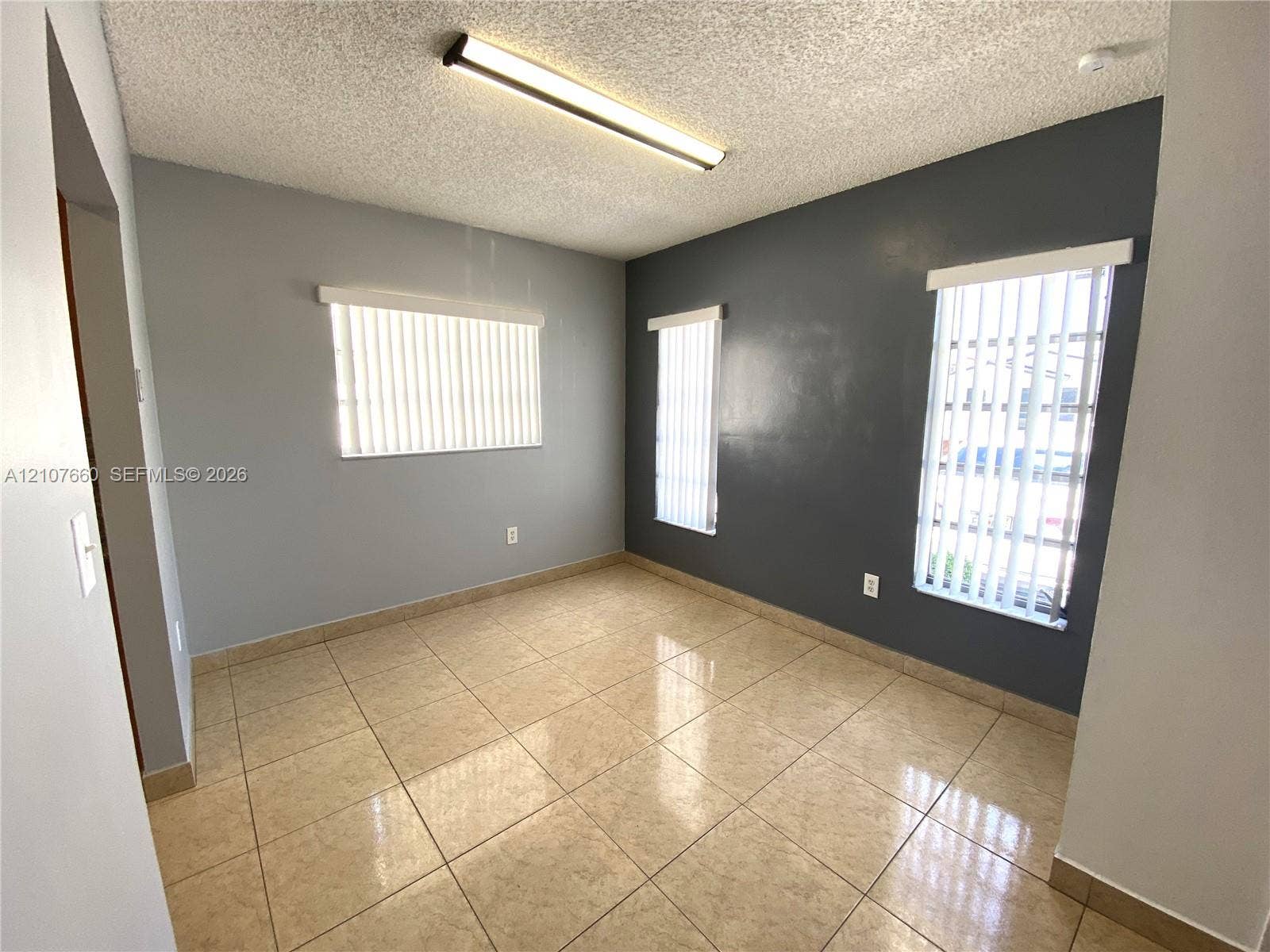 Neutral-toned interior featuring square tile flooring, three windows with vertical blinds, and a linear overhead light fixture