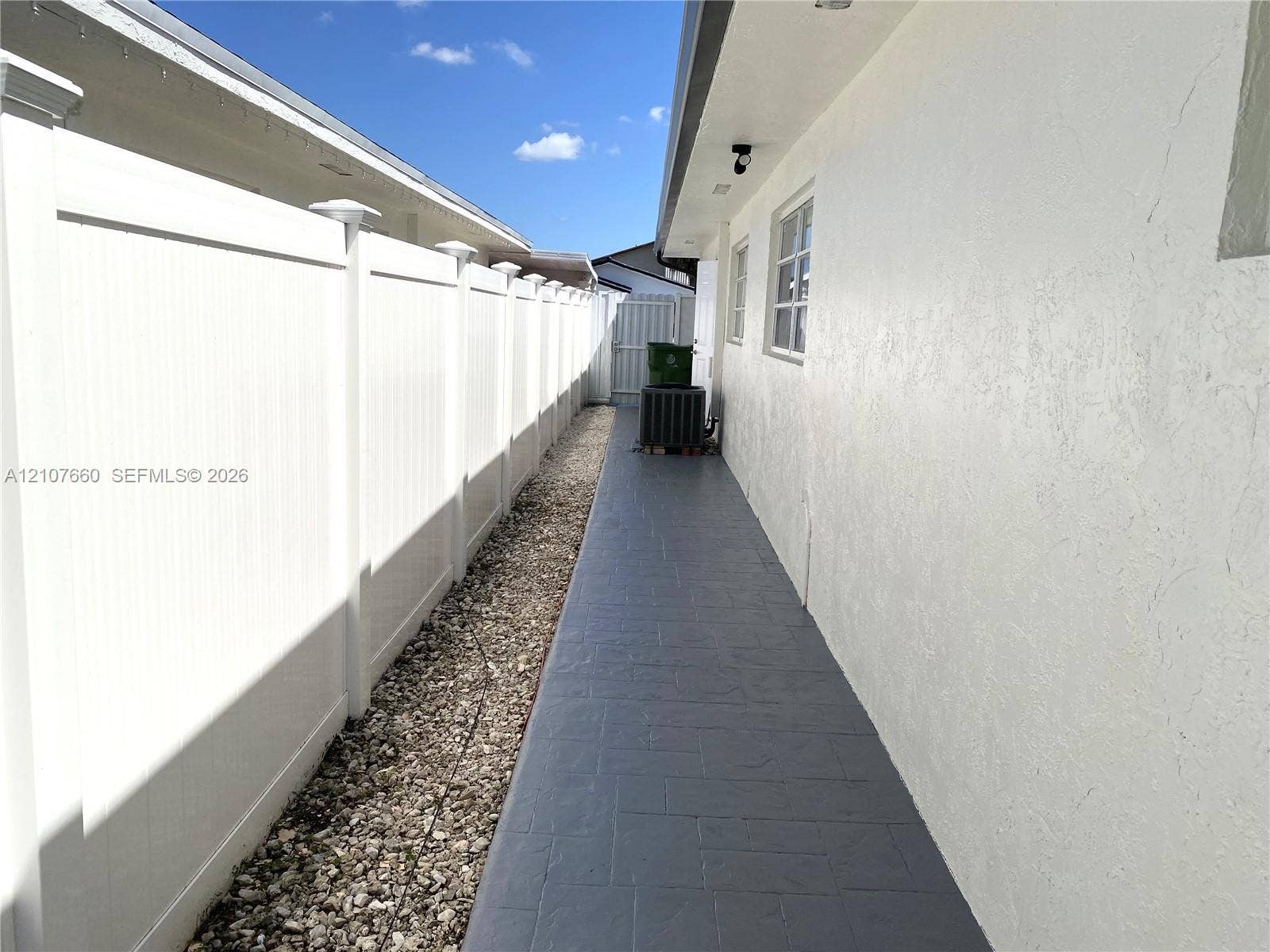 Side yard exterior featuring a white vinyl privacy fence, stamped concrete walkway, and light-toned stucco wall cladding