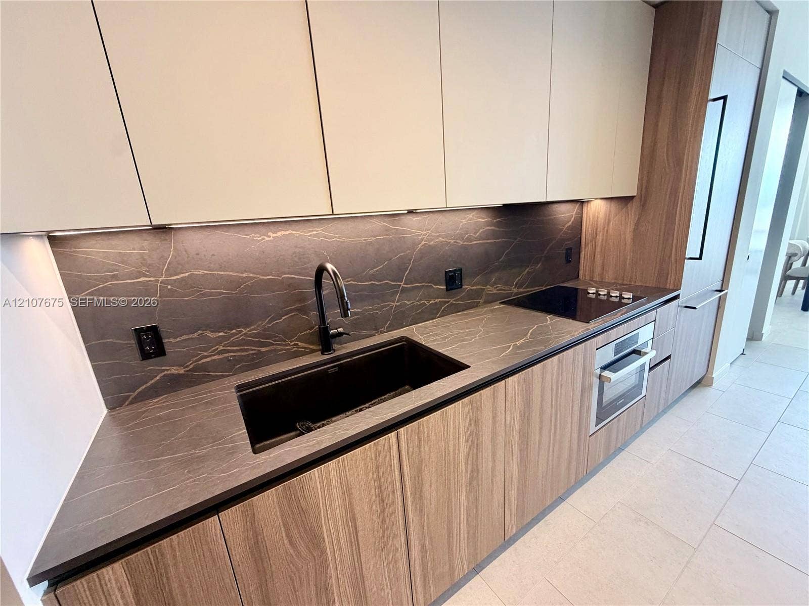 Contemporary kitchen featuring flat-panel wood-finish cabinetry, stone-style countertops and backsplash, an undermount sink with a high-arch faucet, a built-in cooktop, and a stainless steel wall oven