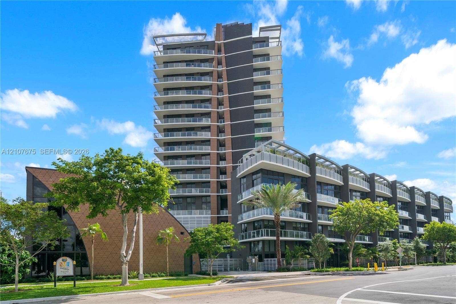 High-rise residential building featuring expansive private balconies with glass railings, multi-story structural architecture, dark exterior paneling, and integrated hardscaping