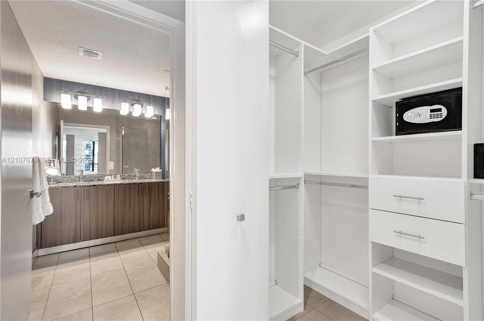 5350 NW 84th Avenue, #1004, Doral, FL 33166 Walk-in closet featuring white built-in shelving, drawers, and hanging rods, accompanied by a bathroom with a dual-sink vanity, stone-finish countertops, tile flooring, and wall-mounted light fixtures