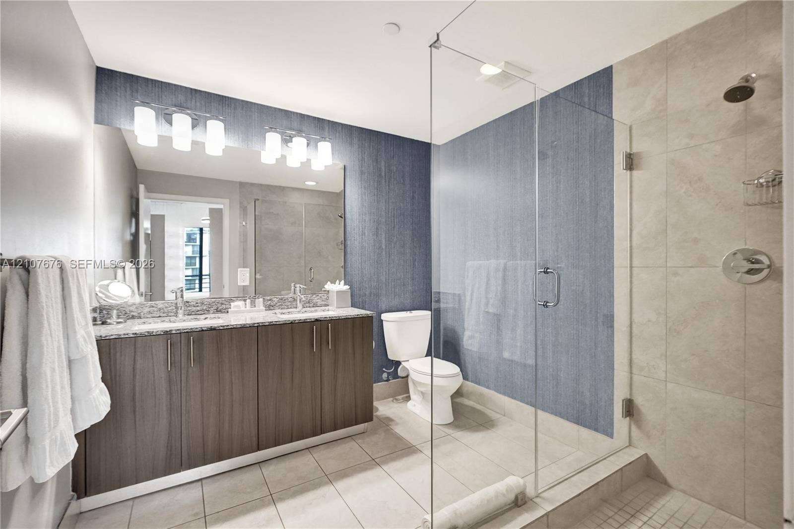 5350 NW 84th Avenue, #1004, Doral, FL 33166 Bathroom featuring a dual-sink vanity with stone countertop, frameless glass-enclosed shower, large-format tile flooring, and integrated wall lighting