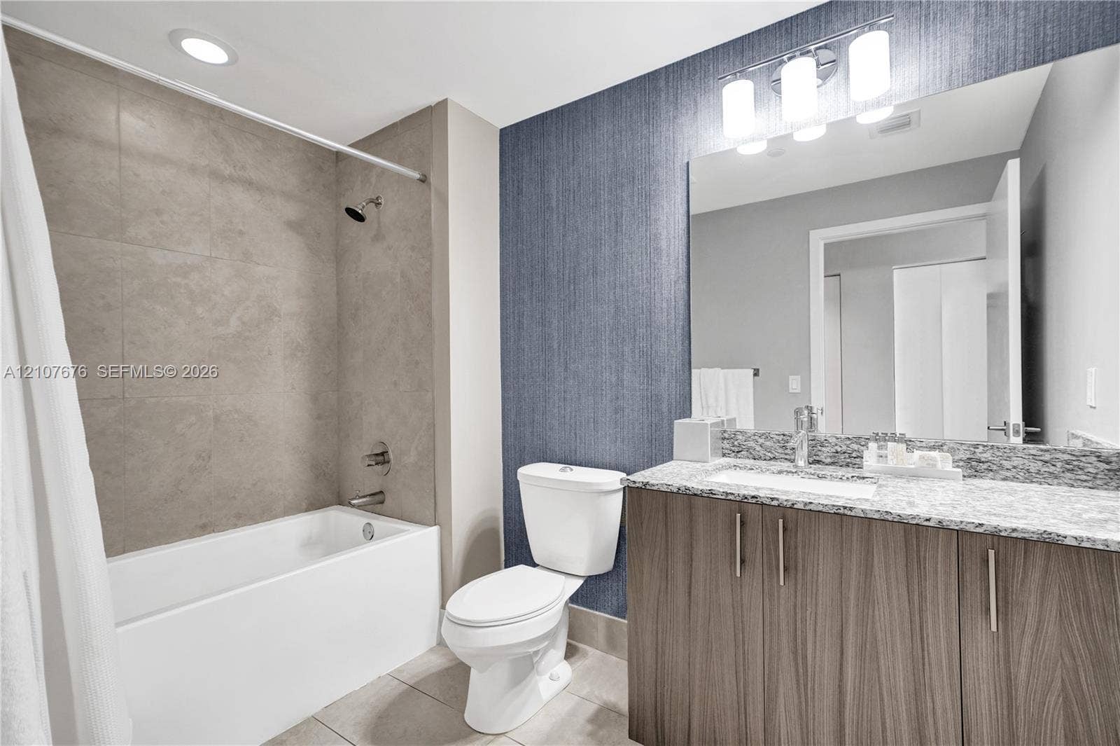 5350 NW 84th Avenue, #1004, Doral, FL 33166 Bathroom featuring a vanity with wood-finish cabinetry, a stone-composite countertop with an integrated sink, textured navy blue wall covering, a large wall-mounted mirror, and a tub-shower combination with floor-to-ceiling tile surround