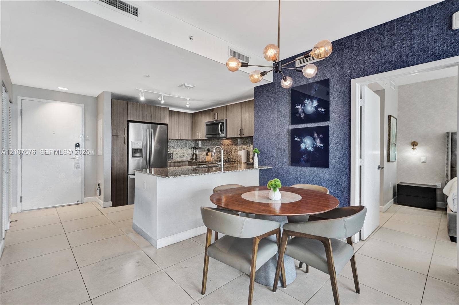 5350 NW 84th Avenue, #1004, Doral, FL 33166 Kitchen featuring stainless steel appliances, stone countertops, and wood-finish cabinetry, complemented by a multi-bulb modern chandelier and a textured accent wall