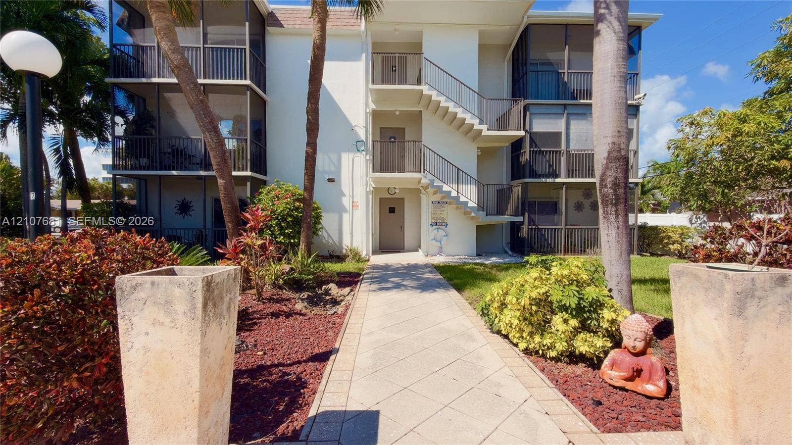 701 NE 1st Court, #202, Hallandale Beach, Hallandale Beach, FL 33009 Three-story residential building exterior featuring screened-in lanais, external staircase, concrete paver walkway, and mature tropical landscaping