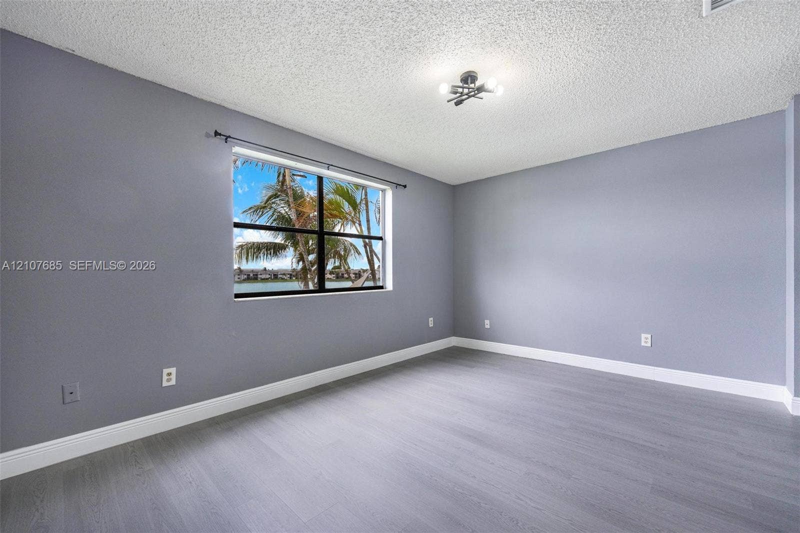 1460 Jefferson Drive, #1460L, Homestead, FL 33034 Room featuring gray-tone walls, wood-finish flooring, a multi-bulb flush-mount light fixture, and a single-pane window with exterior water and palm tree scenery