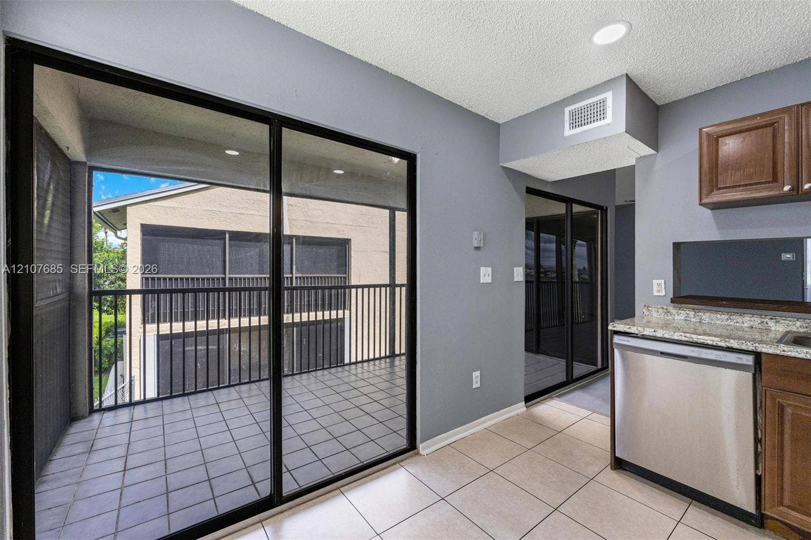 1460 Jefferson Drive, #1460L, Homestead, FL 33034 Sliding glass doors leading to a covered balcony with tiled flooring and metal railing, paired with an interior kitchen featuring stainless steel dishwasher, stone-finish countertops, and wood-finish cabinetry