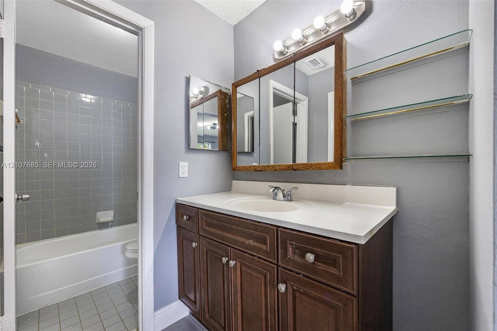1460 Jefferson Drive, #1460L, Homestead, FL 33034 Bathroom featuring a vanity with wood cabinetry, integrated sink, triple-mirror medicine cabinet, four-light vanity fixture, and glass shelving