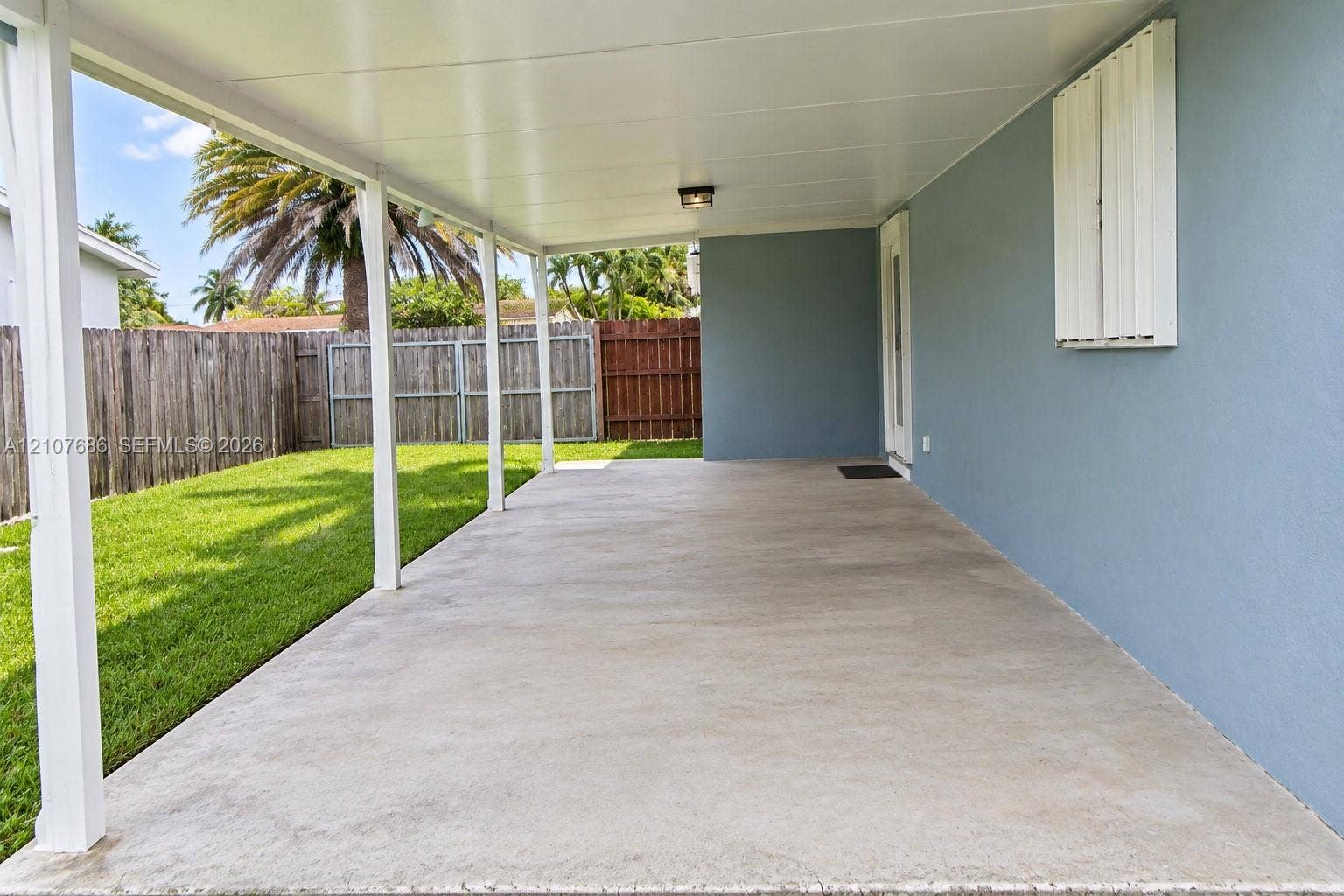 9760 Jamaica Drive, Cutler Bay, FL 33189 Covered concrete patio featuring a solid roof, white support pillars, exterior wall-mounted shutter, and a fenced backyard with lawn