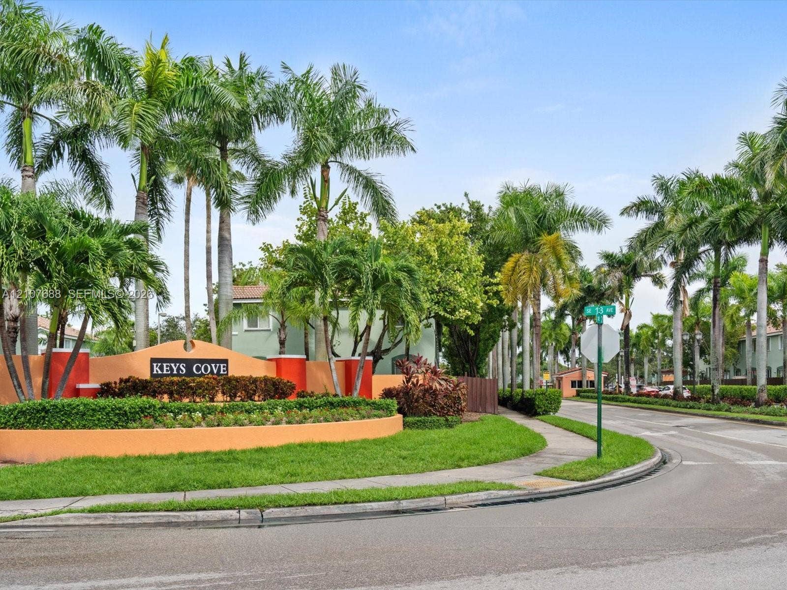 1677 SE 27th Court, #202, Homestead, FL 33035 Community entrance monument with stucco finish, lush landscaping, mature palm trees, manicured lawn, and asphalt paved thoroughfare