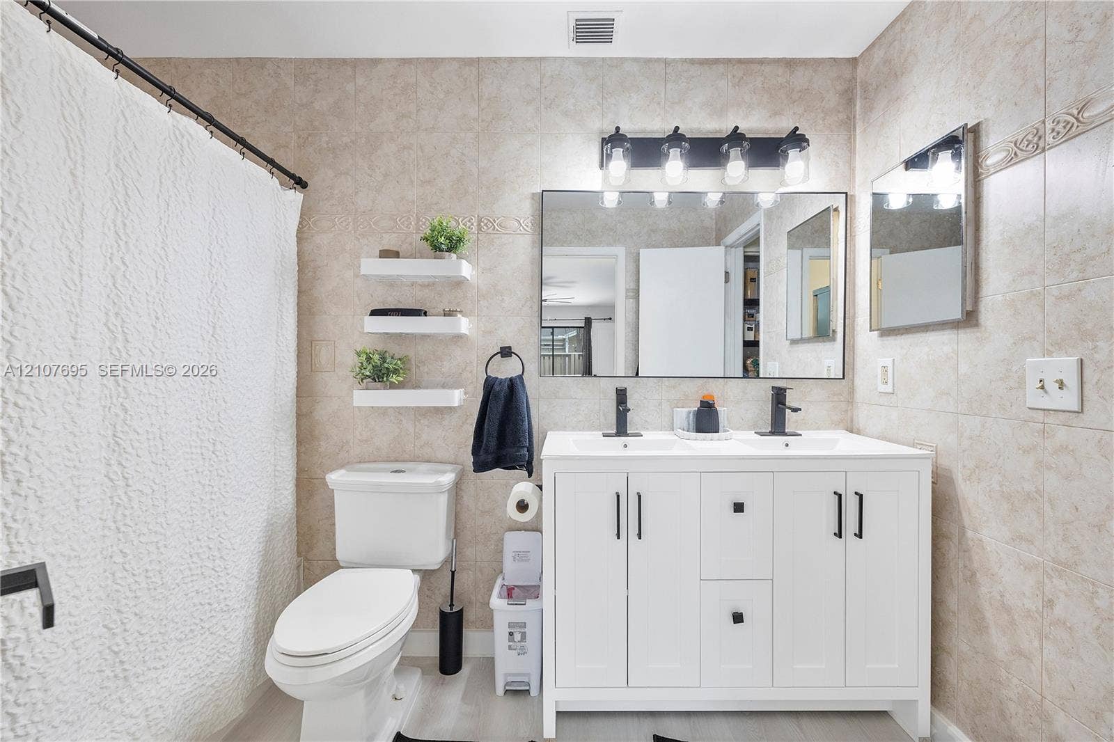 7198 Sportsmans Drive, North Lauderdale, FL 33068 Double-sink vanity with white cabinetry, matte black hardware, and rectangular undermount sinks, complemented by a wide vanity mirror, four-light wall fixture, and neutral-toned wall tiling