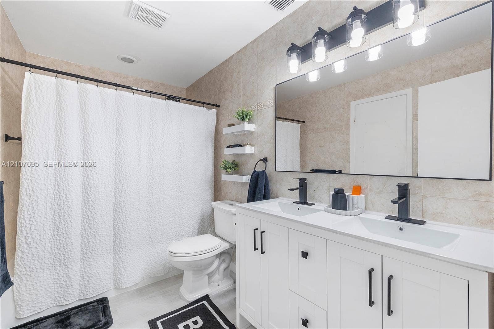 7198 Sportsmans Drive, North Lauderdale, FL 33068 Dual-sink vanity with white cabinetry, matte black hardware, stone-surface countertop, and rectangular undermount sinks, complemented by a large-format mirror with integrated multi-bulb vanity lighting and neutral tile wall cladding