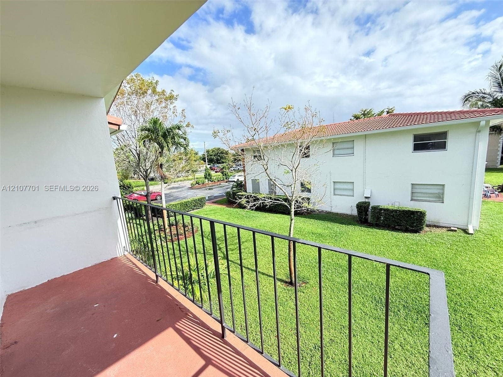 3100 Coral Springs Drive, #2B, Coral Springs, FL 33065 Covered balcony featuring black metal railings and terra-cotta flooring, overlooking a landscaped lawn with mature greenery