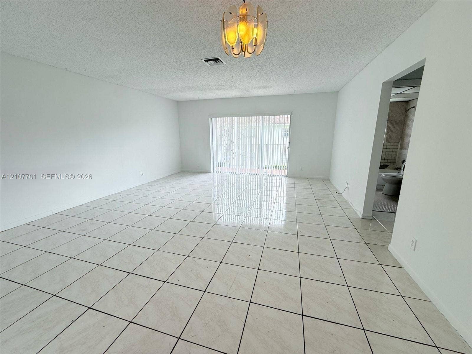 3100 Coral Springs Drive, #2B, Coral Springs, FL 33065 Open-concept area featuring light-toned tile flooring, textured ceiling, glass sliding doors with vertical blinds, and a multi-bulb flush-mount light fixture