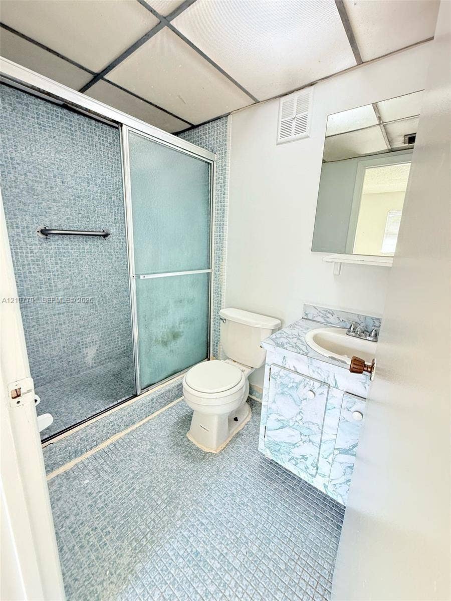 3100 Coral Springs Drive, #2B, Coral Springs, FL 33065 Bathroom featuring a walk-in shower with glass sliding doors, mosaic-tiled flooring and wall surround, a vanity with a patterned stone-finish countertop, and a wall-mounted mirror
