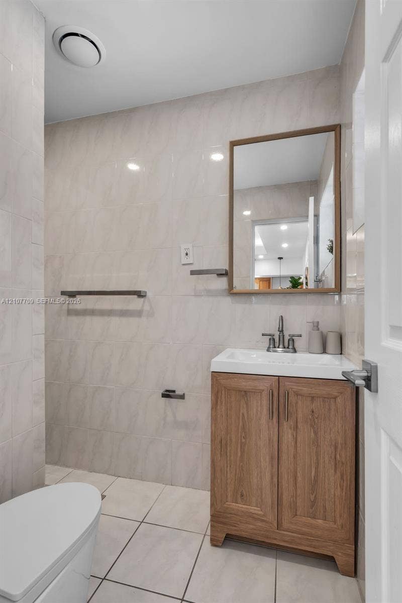 Bathroom featuring a wood-finish vanity with a rectangular white sink, metallic faucet, framed wall mirror, and floor-to-ceiling tile work