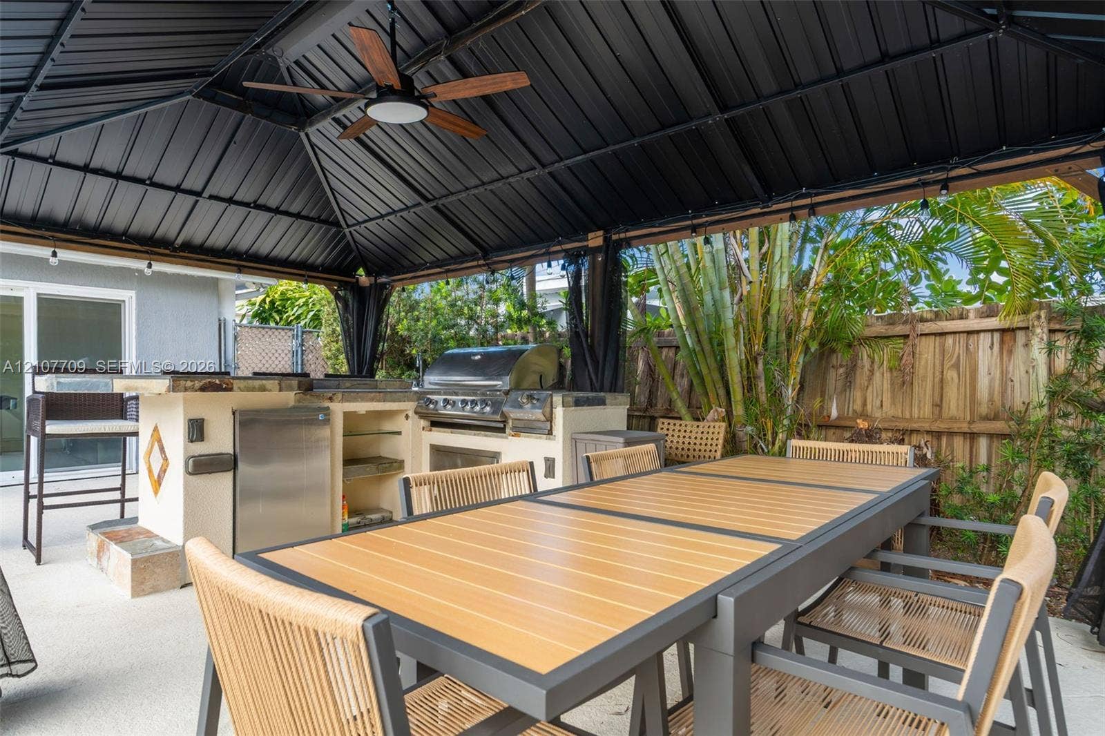Covered outdoor kitchen featuring a metal roof structure, built-in stainless steel grill, integrated mini-refrigerator, stone-finish base, and overhead ceiling fan