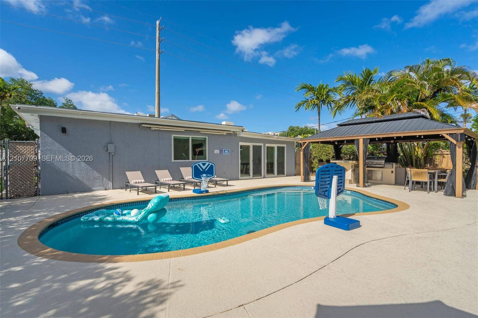 Inground swimming pool with a paved deck, accompanied by a covered outdoor gazebo featuring a built-in kitchen station