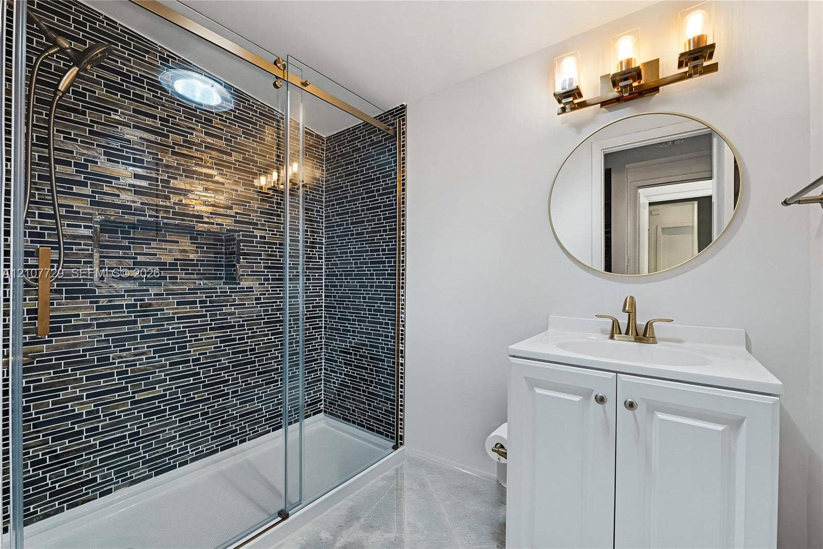 Bathroom featuring a walk-in shower with glass sliding doors and multi-tonal linear tile surround, brass-finish fixtures, white vanity cabinet with an integrated basin, and a circular wall-mounted mirror with a matching brass-finish light fixture