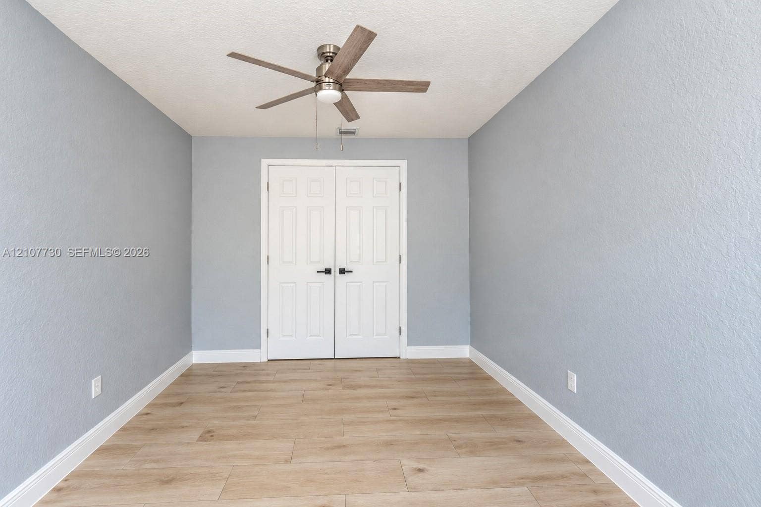 15606 SW 52nd Court, Miramar, FL 33027 Room featuring wood-finish tile flooring, light blue textured walls, dual-panel closet doors with matte black hardware, and a ceiling fan with integrated lighting