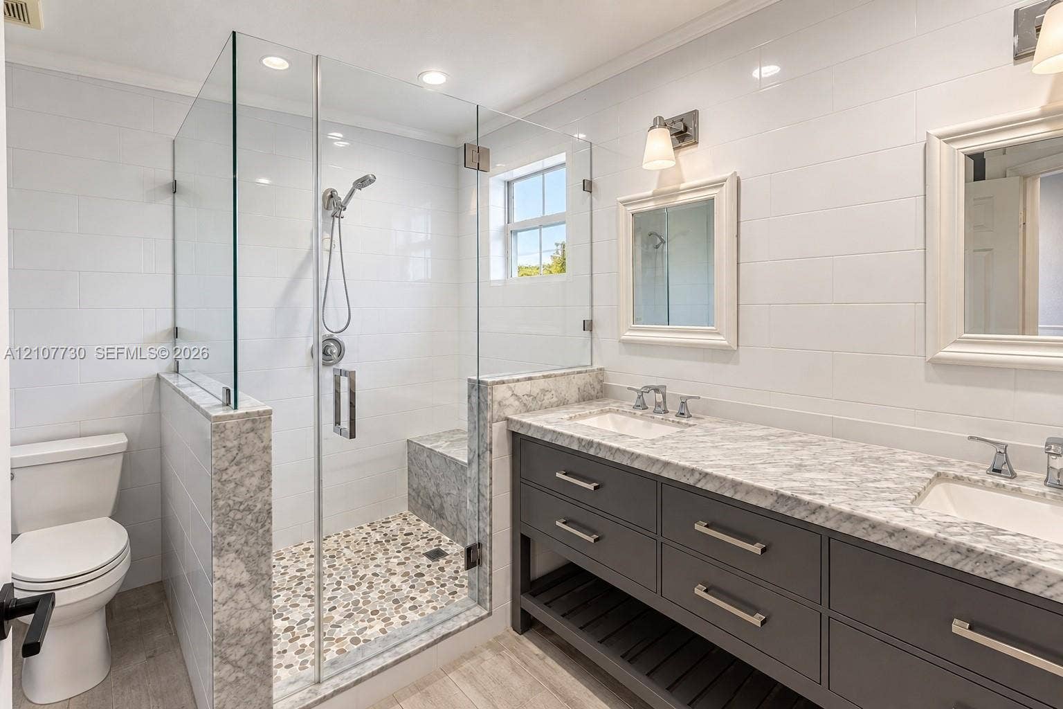 15606 SW 52nd Court, Miramar, FL 33027 Double vanity with stone countertop, glass-enclosed walk-in shower featuring an integrated bench seat, pebble-style shower flooring, and wall-mounted light fixtures