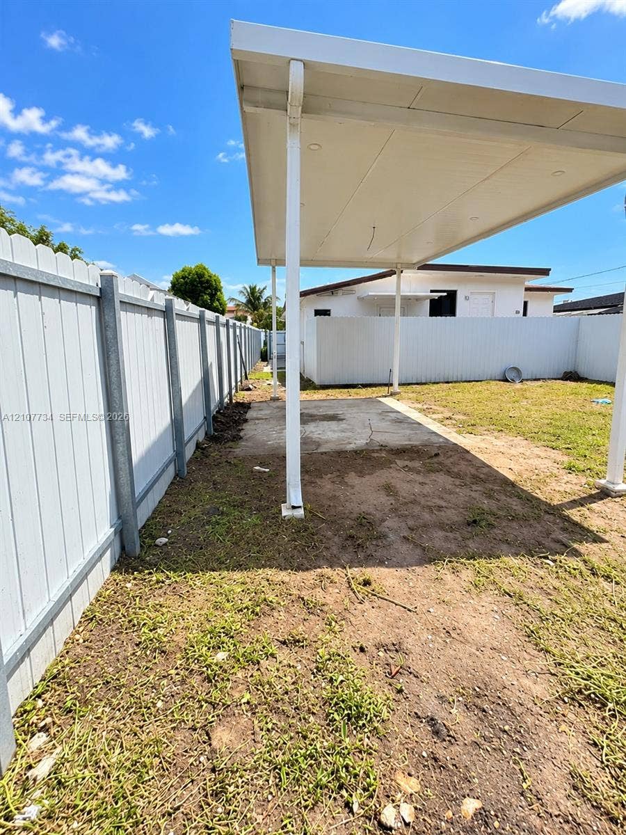 Covered patio featuring a concrete slab and white metal support posts, enclosed by a white vinyl perimeter fence