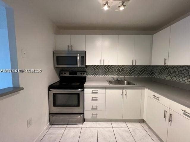 Kitchen featuring white cabinetry, stainless steel microwave and range, solid-surface countertops, mosaic tile backsplash, and light-toned tile flooring