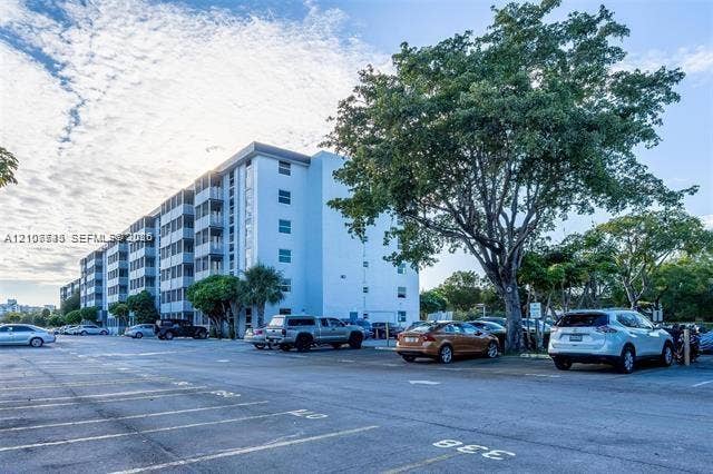 Multi-story residential building featuring a light-toned stucco exterior, vertical structural columns, and an expansive asphalt parking area with designated numbered spaces