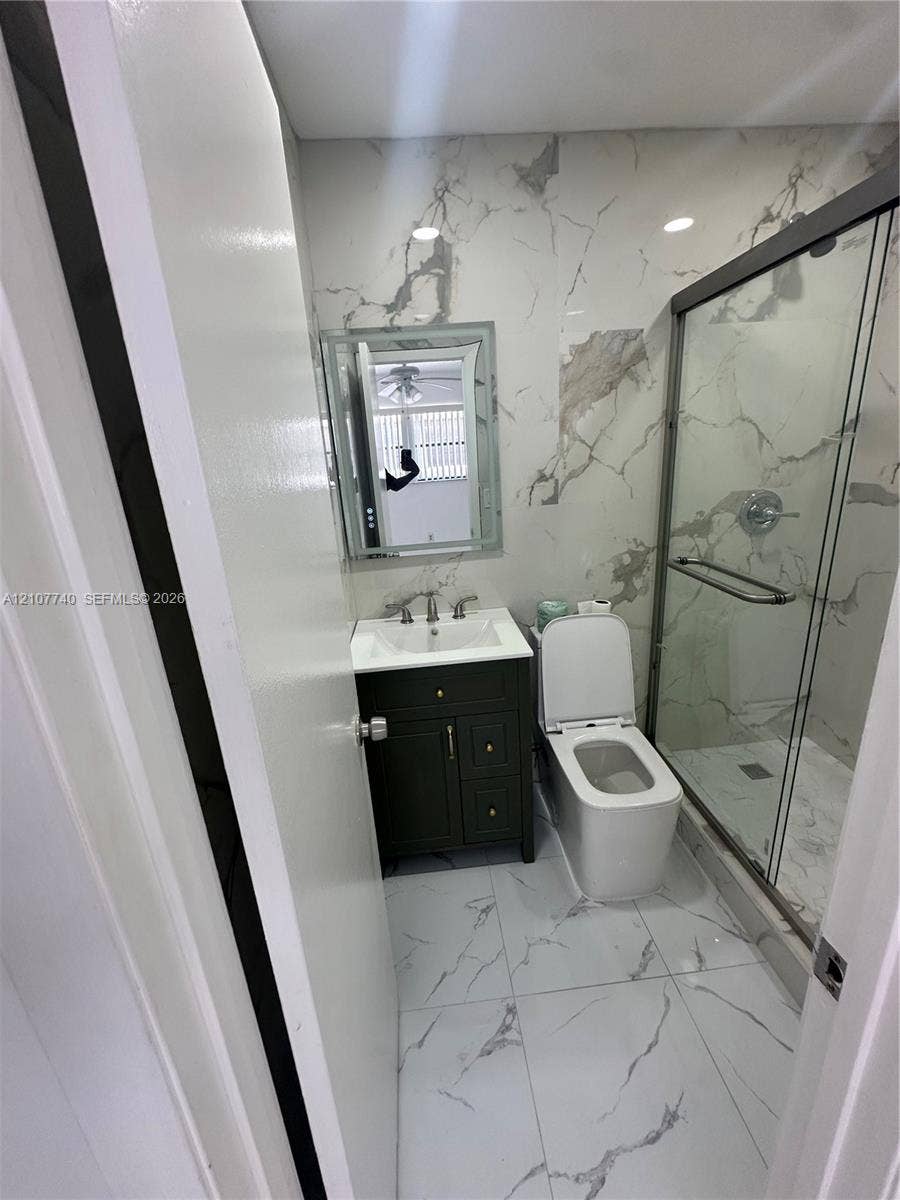 Full-height stone-patterned wall tiling, glass-enclosed shower with metallic hardware, dark-finish wood vanity with integrated sink, and large-format porcelain floor tile