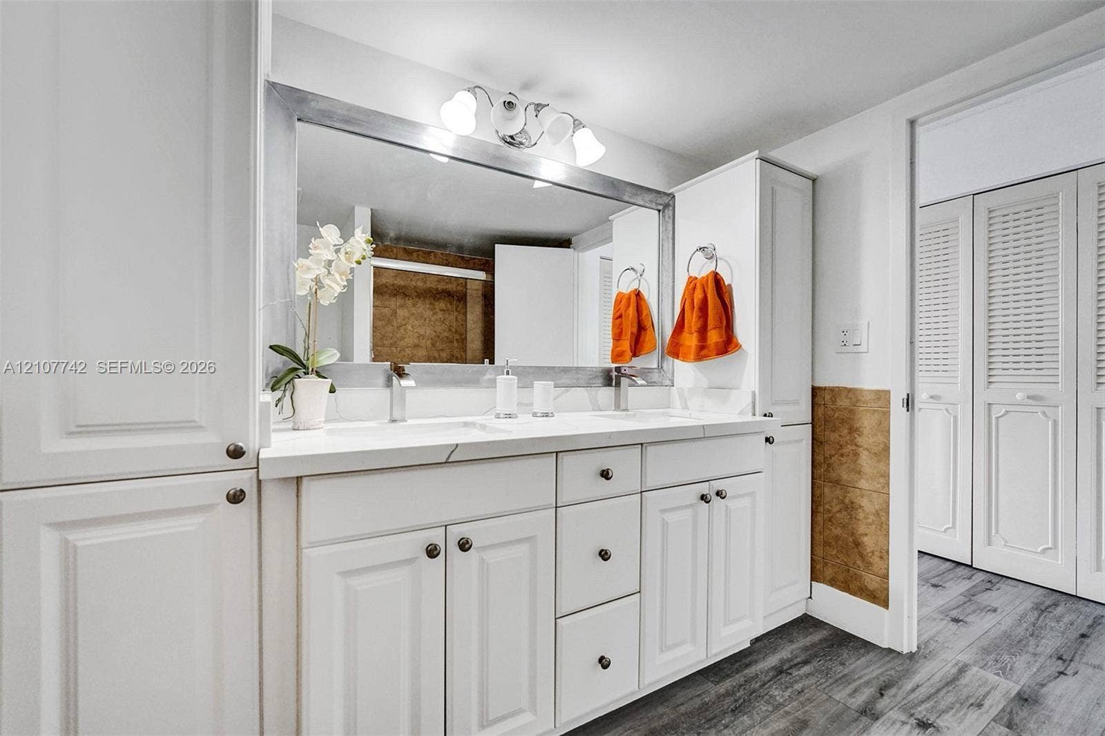 100 Golden Isles Drive, #112, Hallandale Beach, Hallandale Beach, FL 33009 Dual-sink vanity with white cabinetry, solid surface countertop, large framed wall mirror, multi-bulb overhead lighting, and white louvered closet doors