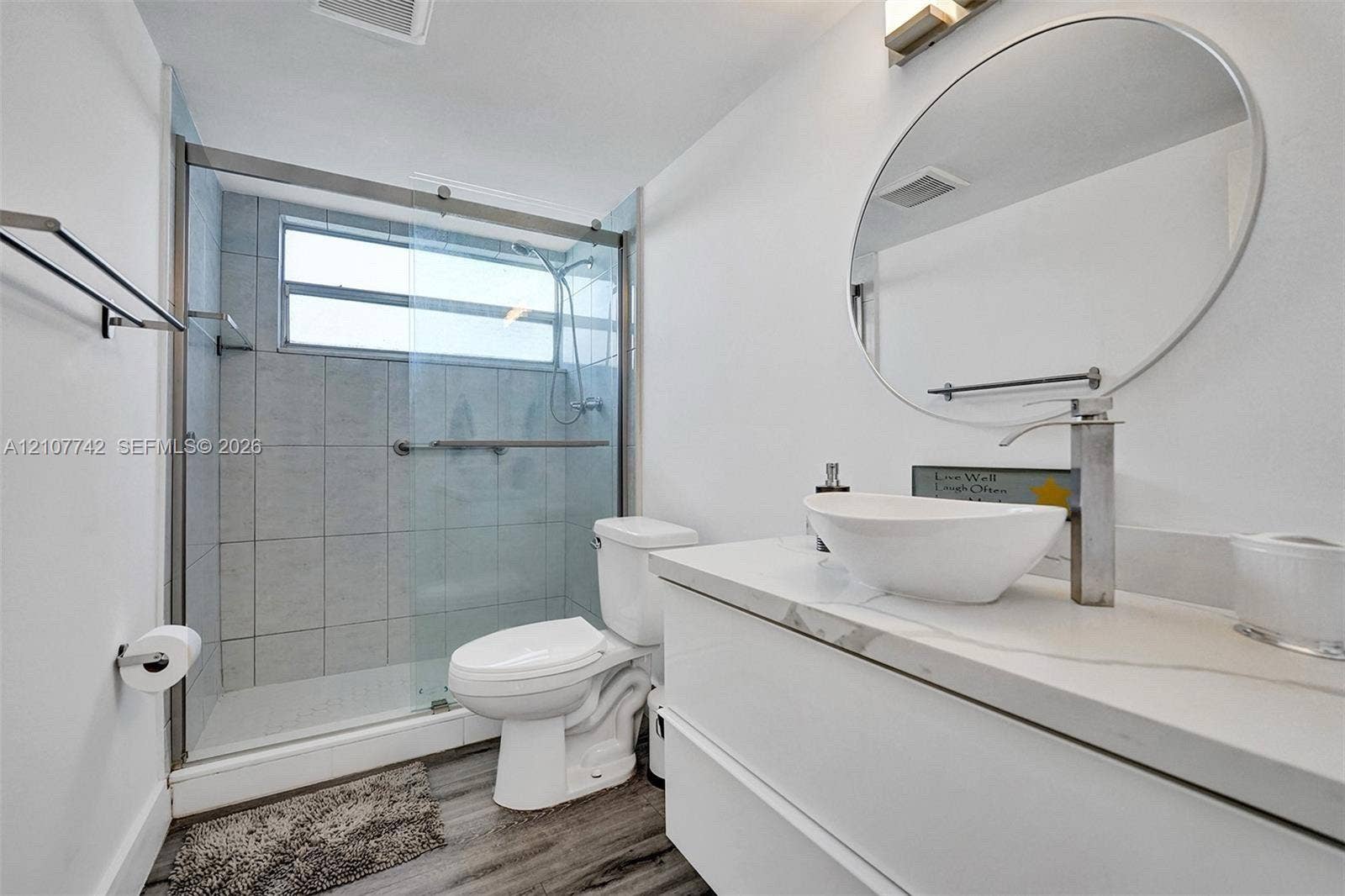 100 Golden Isles Drive, #112, Hallandale Beach, Hallandale Beach, FL 33009 Modern bathroom featuring a floating vanity with a white countertop and vessel sink, circular mirror, and walk-in shower with a glass enclosure and tiled surround