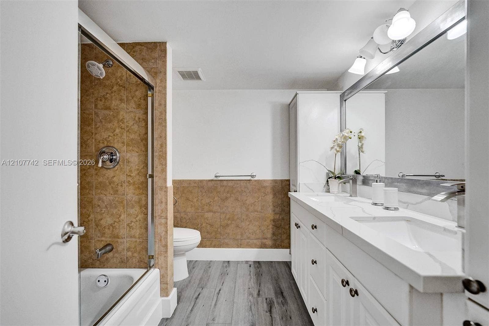 100 Golden Isles Drive, #112, Hallandale Beach, Hallandale Beach, FL 33009 Bathroom featuring a dual-sink vanity with a solid-surface countertop, full-width mirror, shower-tub combination with tiled surround, and wood-finish flooring