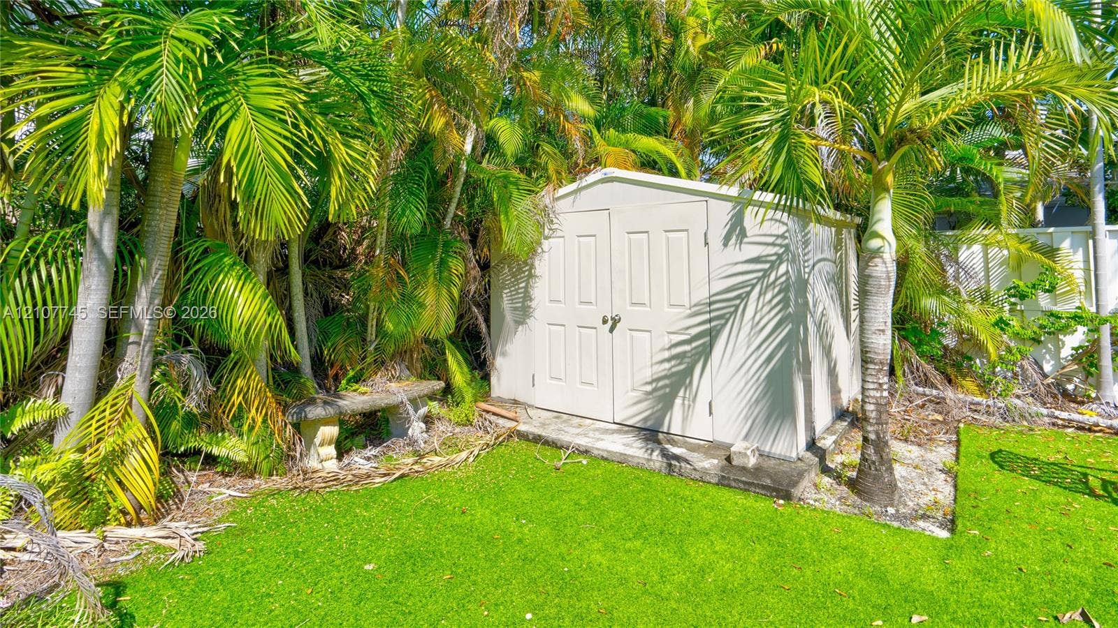 Exterior storage shed featuring double doors and a concrete foundation pad, complemented by mature palm trees and stone bench seating