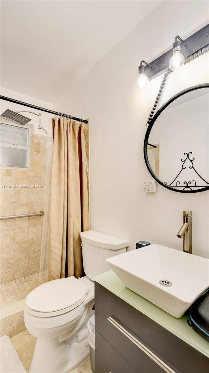 Bathroom featuring a vessel sink with a modern faucet, dark cabinetry, a round decorative mirror, and a walk-in tiled shower with a rainfall showerhead