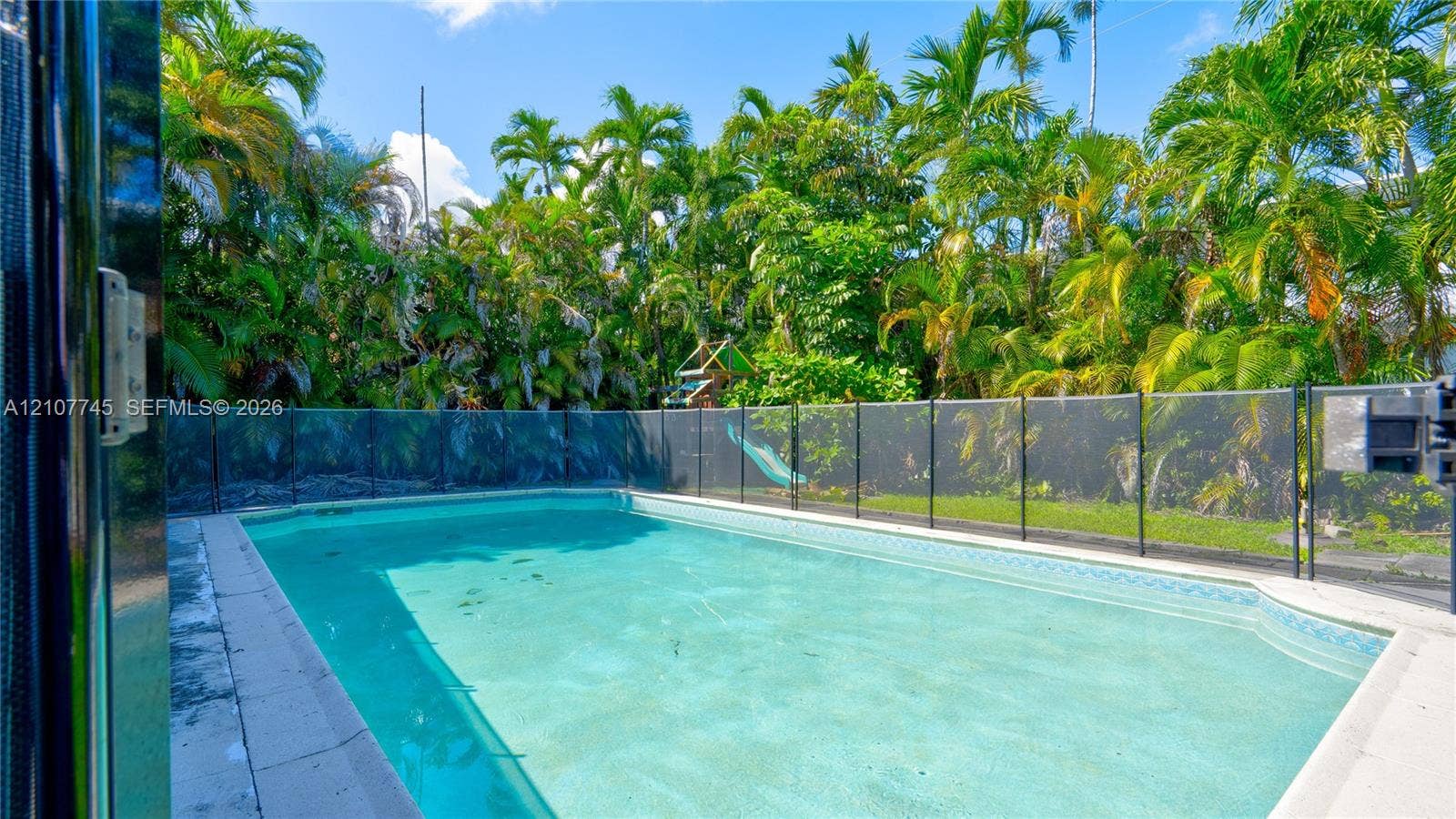 In-ground swimming pool featuring a perimeter safety fence, concrete pool deck, and mature palm tree landscaping