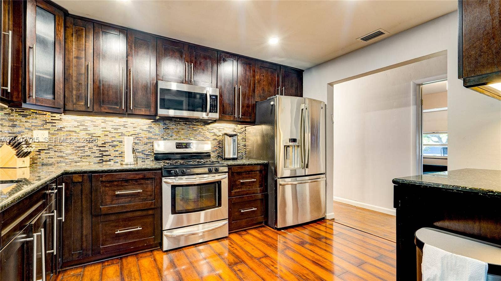 Kitchen featuring dark wood cabinetry, stainless steel appliances, multi-tonal mosaic tile backsplash, stone-finish countertops, and wood-finish flooring