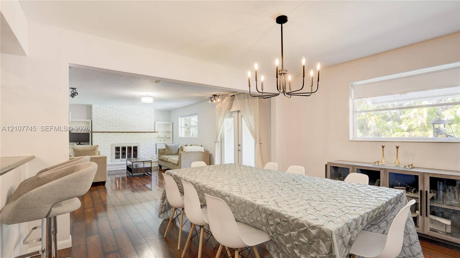 Dining area featuring wood-finish flooring, a multi-arm candle-style chandelier, and a large window, with a painted brick fireplace visible in the background