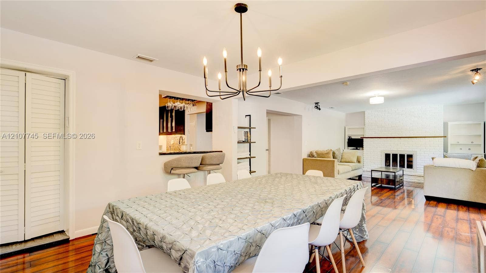 Open-concept interior featuring wood-finish flooring, a multi-arm candle-style chandelier, a pass-through bar with built-in counter, and a painted brick fireplace
