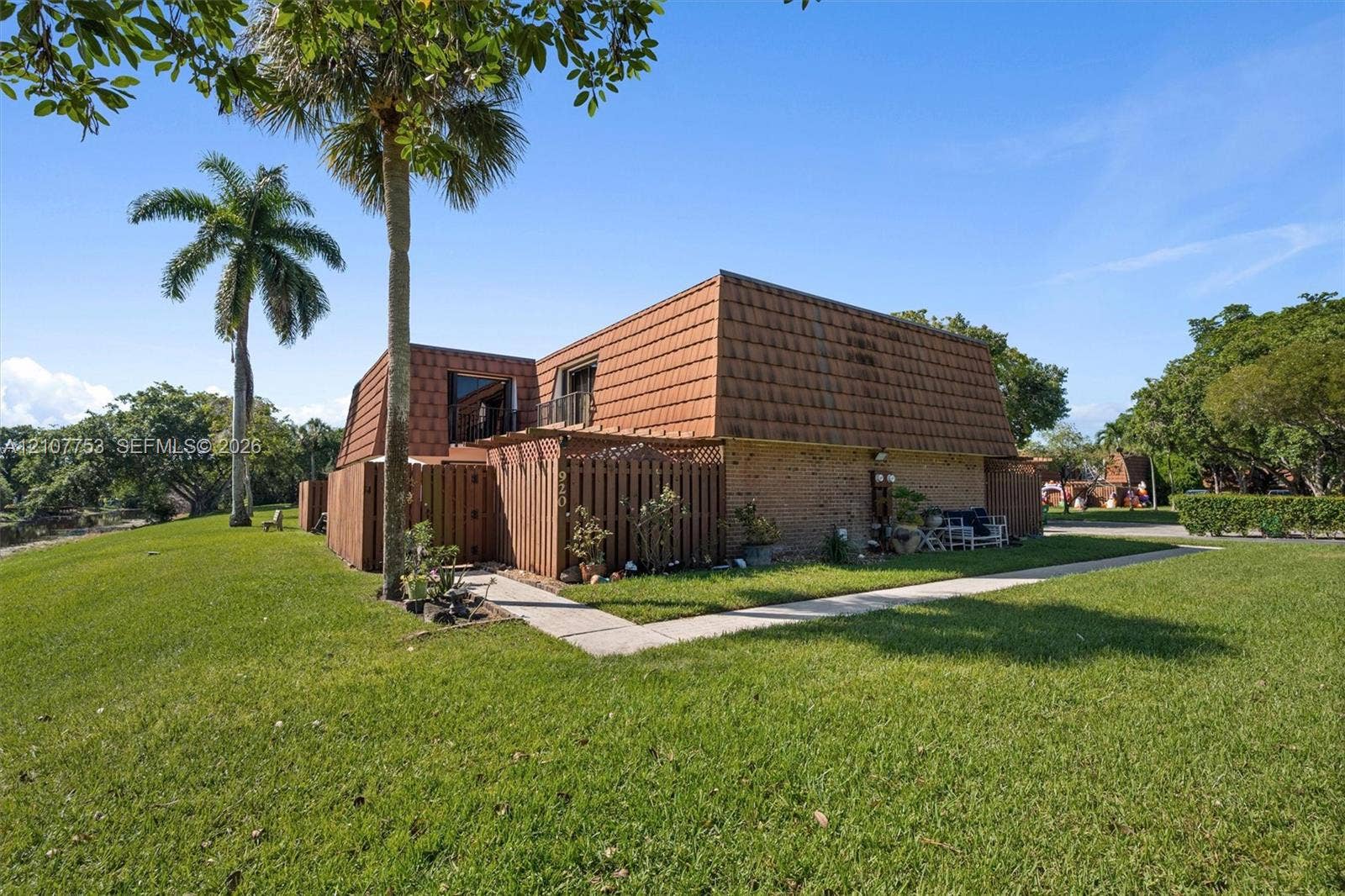 Exterior featuring a mansard roof, brick masonry, wood-lattice privacy screening, and concrete walkway set within a manicured lawn