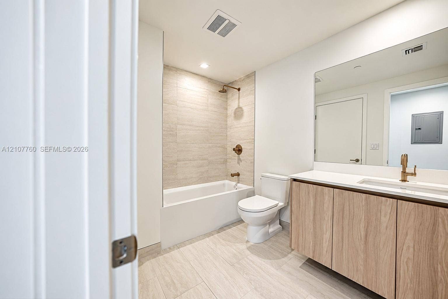 Bathroom featuring a wood-finish floating vanity, neutral-toned tile flooring, large wall-mounted mirror, and a tub with tiled surround and bronze-tone fixtures