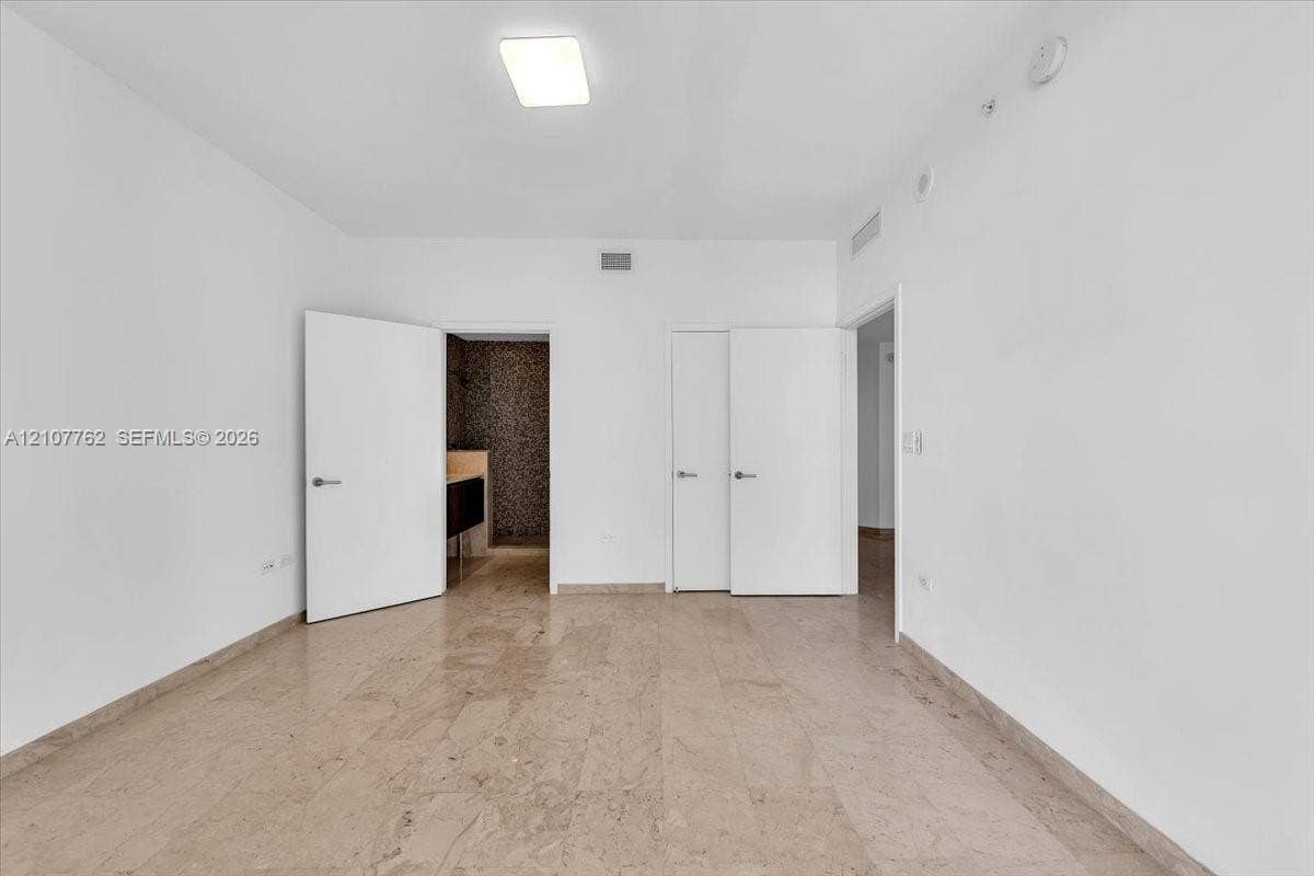 Spacious room featuring neutral stone flooring, white walls, and multiple closet doors with brushed metal hardware