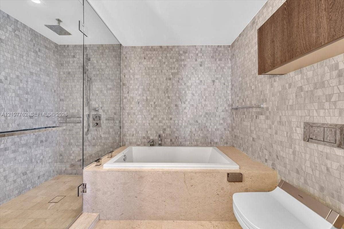 Soaking tub with stone surround, walk-in shower with frameless glass enclosure, rainfall showerhead, wall-mounted toilet, and mosaic tile wall accents