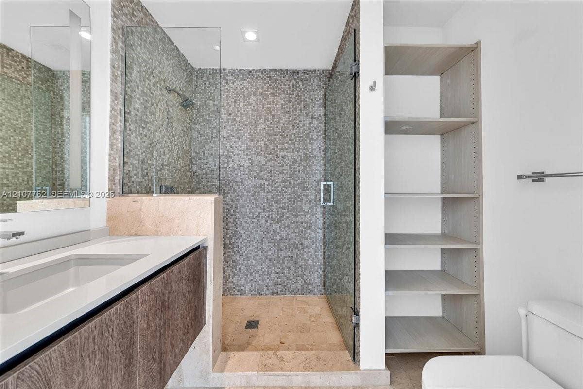 Floating vanity with a smooth-surface countertop, shower enclosed by frameless glass featuring floor-to-ceiling mosaic tile, and built-in wood-finish shelving