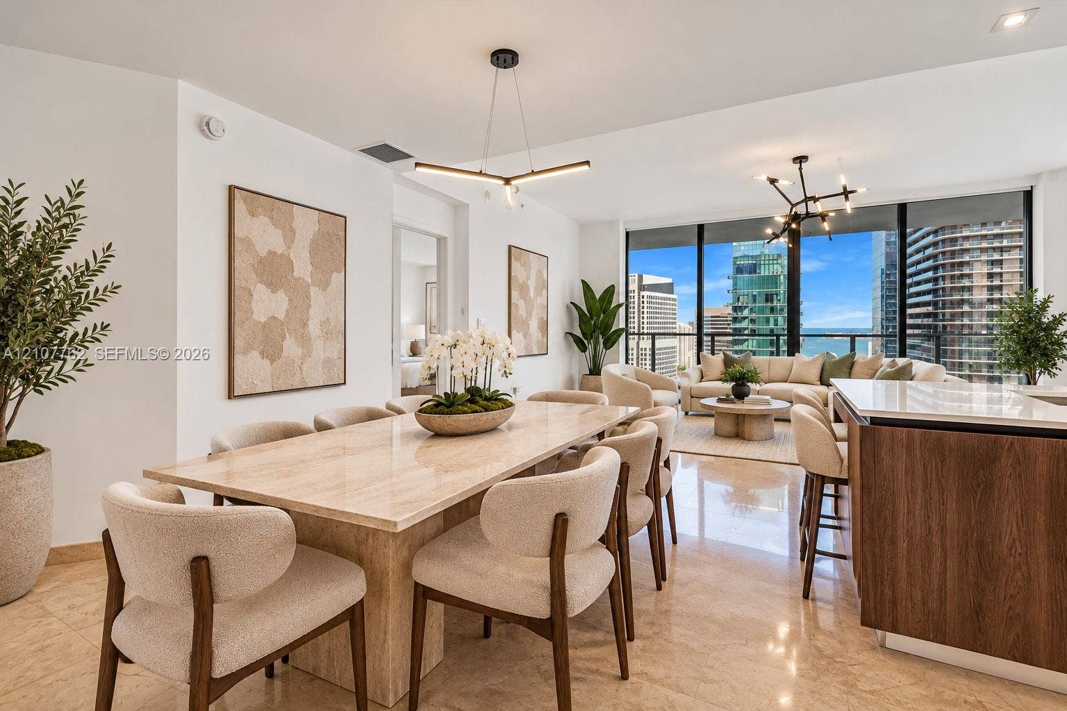 Open-concept interior featuring floor-to-ceiling windows, polished stone flooring, modern linear chandeliers, and an island with a wood-finish base and light-toned stone countertop