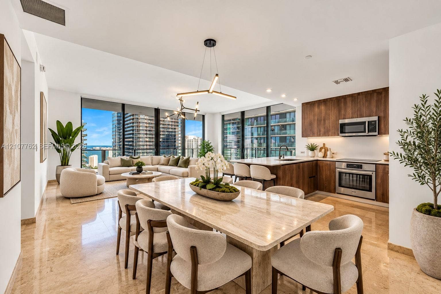 Open-concept interior featuring floor-to-ceiling windows, natural stone flooring, linear modern pendant lighting, and wood-finish cabinetry with built-in stainless steel appliances